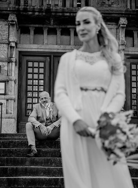 Braut schaut leicht lächelnd in die Kamera im Hintergrund sitzt der Bräutigam auf Treppenstufen und schaut in die Kamera. Die Schärfe liegt dabei auf dem Bräutigam. Aufgenommen vor der Kieler Oper.