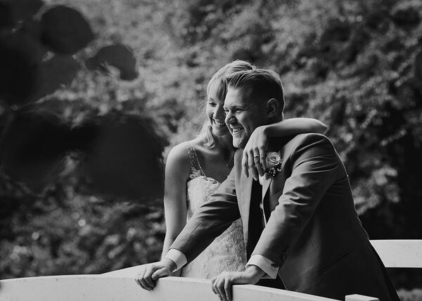 Brautpaar steht auf Brücke. Bräutigam stützt sich auf dem Geländer ab und die Braut umarmt ihn.