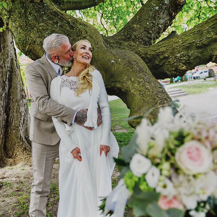Bräutigam umarmt Braut und küsst sie am Hals. Sie stehen an einem Baum. Im Vordergrund liegt der Brautstrauß in der Unschärfe.