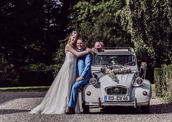 Bräutigam sitzt auf Kotflügel eines Autos (Ente). Die Braut steht neben ihm und umarmt ihn.
