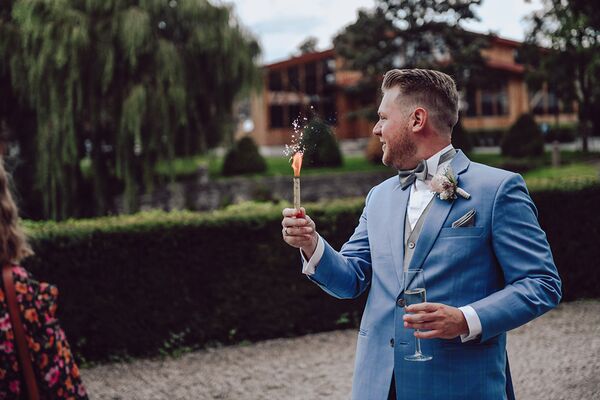 Bräutigam steht auf dem Vorplatz und hält eine Fontäne in der einen Hand und in der anderen ein Sektglas.