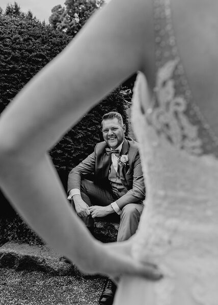 Im Vordergrund der angewinkelte Arm der Braut- unscharf. Der Blick geht durch den angewinkelten Arm hindurch auf dem Bräutigam der auf einer Treppe sitzt.