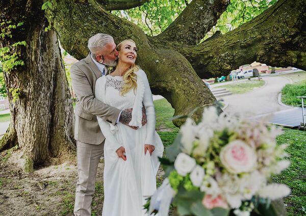 Bräutigam umarmt Braut und küsst sie am Hals. Sie stehen an einem Baum. Im Vordergrund liegt der Brautstrauß in der Unschärfe.