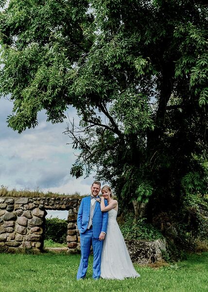 Brautpaar steht auf Wiese vor einem großen Baum. Das Paar ist dabei im Verhältnis zum Baum eher klein auf dem Bild. Die Braut lehnt sich an der Schulter des Bräutigams an.