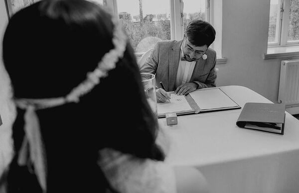 Bräutigam unterzeichnet die Eheurkunde. Braut ist im Vordergrund unscharf und schaut zum Bräutigam.