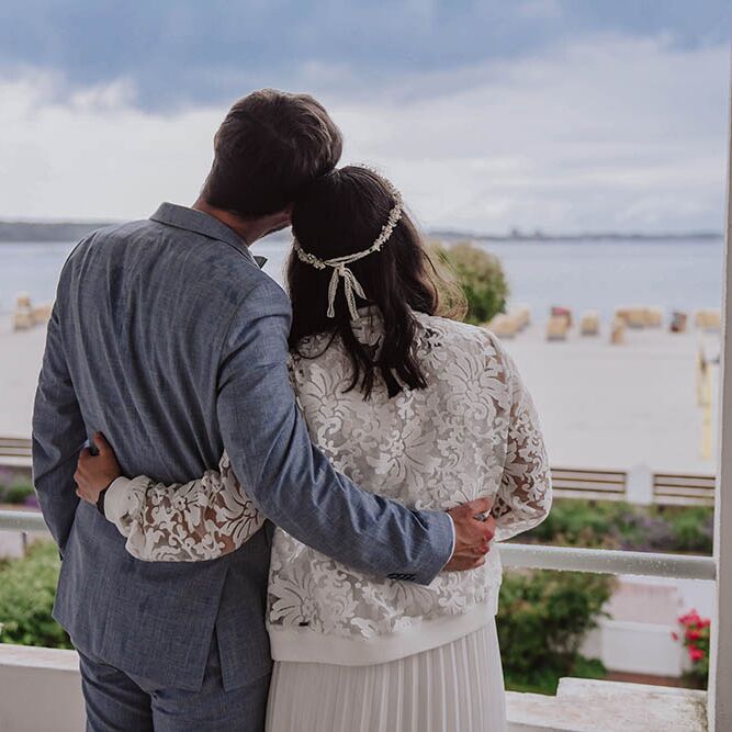 Brautpaar steht auf Balkon und schaut auf den Strand in Laboe und auf's Meer hinaus.